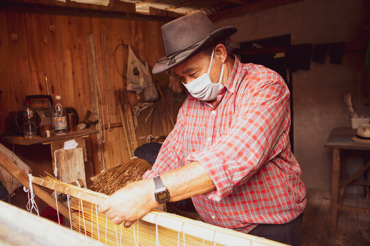 Traditional handicraft mat made of dry reed in a local community of Garopaba, Brazil. Artisan working at his wooden workshop. Craftsman wearing face-mask during covid-19 pandemic.