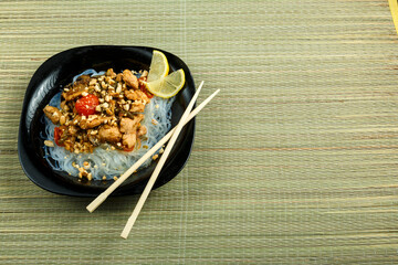 plate with funchose and chicken with vegetables in pad Thai sauce next to chopsticks on a bamboo mat.