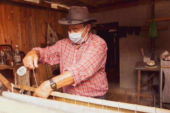 Traditional handicraft mat made of dry reed in a local community of Garopaba, Brazil. Artisan working at his wooden workshop. Craftsman wearing face-mask during covid-19 pandemic.