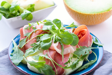 Traditional Italian or Spanish appetizer fresh melon with prosciutto or jamon and green herbs on blue plate and white table, top view, light mediterranean cuisine dish