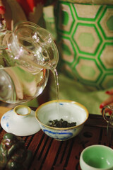 A girl in a green kimono pours water from a transparent teapot into a white bowl with tea on a wooden tray
