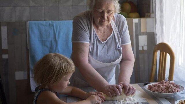 Funny blond boy helps beloved  grandmother in kitchen. elderly woman rolls pastry for pasties and puts minced meat. teenager touches dough. Kid and granny are happy together. shallow depth of field 