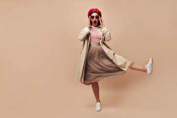 Woman in eyeglasses surprised looks into camera on beige background. Stylish girl in sunglasses in shape of heart and in long skirt raises her leg..