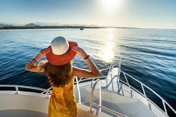 Young woman with long hair wearing yellow dress and straw hat standing on white yacht deck enjoying...