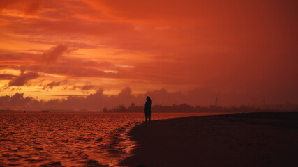 Fototapeta premium Woman walks alone on the beach at the colorful sunset after the rain. Legs in the white shoes. Red orange, crimson purple colours.