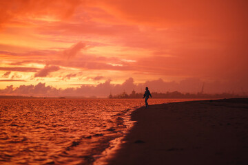 Fototapeta premium Woman walks alone on the beach at the colorful sunset after the rain. Legs in the white shoes. Red orange, crimson purple colours.