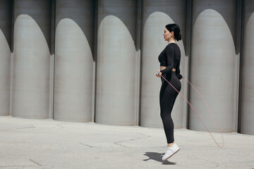 Profile of a sports woman who jumps on a rope in the street in a park against a concrete wall - obrazy, fototapety, plakaty