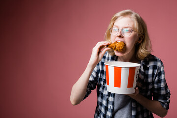 Portrait of a blonde girl in glasses who holds a bucket of fast food in her hand and eats a chicken...