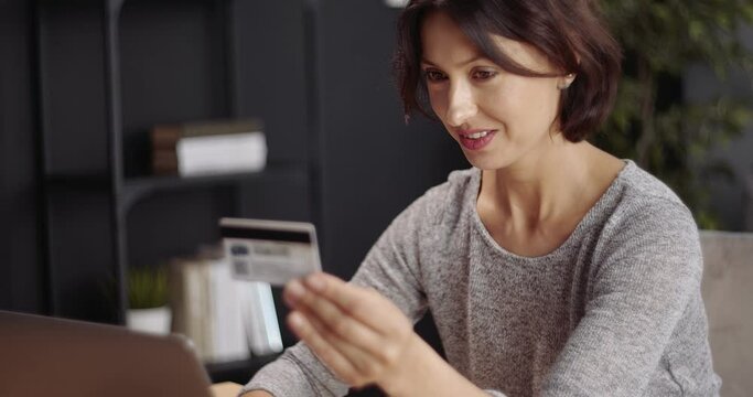 Smiling mature woman in domestic outfit holding credit card and typing all information on laptop for online purchases. Attractive lady ordering goods through internet while staying at home.