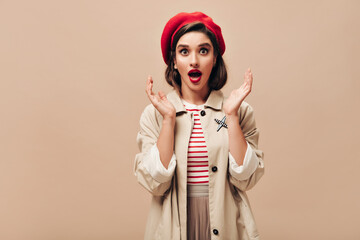 Shocked lady in red hat and beige trench looks into camera. Brown-eyed girl with bright lips in stylish outfit poses on isolated background.. - obrazy, fototapety, plakaty