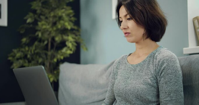 Charming mature woman in casual wear relaxing on grey sofa and browsing internet on modern laptop. Concept of people, technology and leisure time.