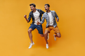Fototapeta na wymiar Excited emotional men in stylish colorful shorts and checkered shirts rejoice and jump on orange background.