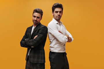 Serious men in suits and white shirts cross arms and look at camera. Young guys in classical style outfits pose on orange background.