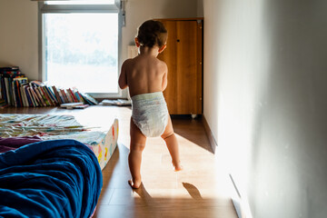 Baby in diapers learning to walk in her bedroom barefoot. - obrazy, fototapety, plakaty