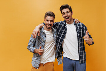 Excited brunet men in checkered shirts, colorful pants and white t-shirts smile, show thumbs up and pose on orange background. - obrazy, fototapety, plakaty