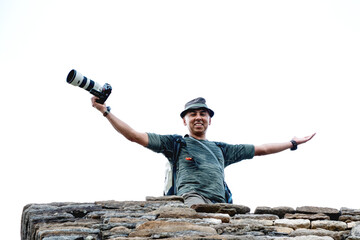 Photographer smiles and looks at camera with camera in hand and open arms over stone wall and cloudy sky 