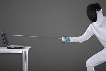 Man in fencing suit aiming fencing foil at a laptop