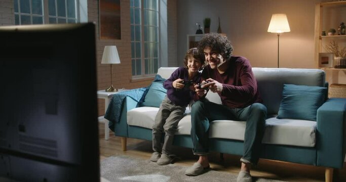 Funny asian siblings with curly hair are having fun at home, playing video games in front of tv, authentic father and son enjoying their hobby 4k footage