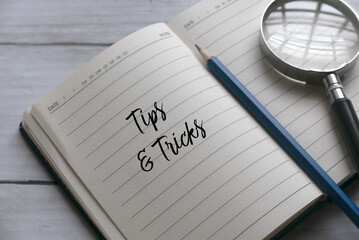 Selective focus of magnifying glass,pencil and notebook written with Tips and Tricks on white wooden background.