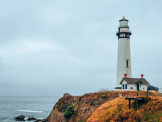 Naklejka premium Pigeon point Lighthouse in Pescadero. White lighthouse on the Pacific coast. The tragic dark sky. Colorful flowers. Fall, summer. Free space for text. California, USA