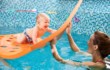 Snapshot of a very happy and playful swimming class with instructor. Cheerful baby boy is crawling on a floating mat with holes and playing with a caregiver