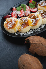 Closeup of fried curd fritters with jam, coconut flakes and strawberries, vertical shot on a black stone background