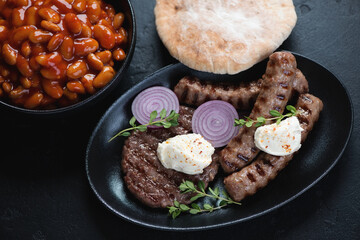 Grilled serbian pljeskavica and cevapi sausages with prebranac and pita over black stone surface, horizontal shot, selective focus