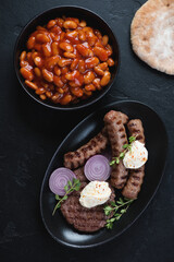 Grilled balkan meat patty or pljeskavica with cevapcici or skinless beef sausages and prebranac, above view on a black stone background, vertical shot