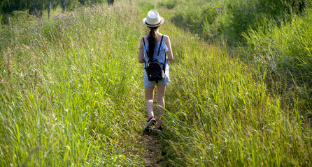 Beautiful  girl tourist with backpack going camping in summer forest. Forest Adventure Travel Remote Relax Concept. The girl walks along the path in the plants in a light blue suit and hat