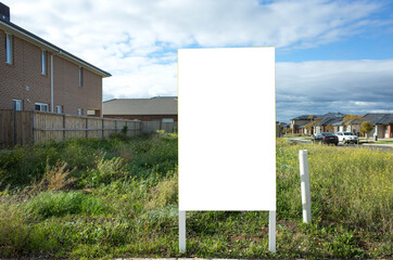 Blank white empty mockup template of a real estate sign at front of a lot of vacant land on a suburban neighbourhood street with some residential houses at the background.