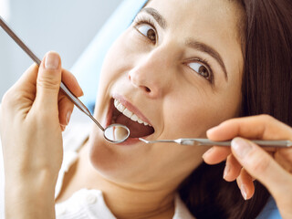 Smiling brunette woman being examined by dentist at dental clinic. Hands of a doctor holding dental instruments near patient's mouth. Healthy teeth and medicine concept