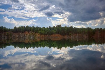 Beautiful summer landscape, forest trees are reflected in calm river water against a background of blue sky and white clouds.