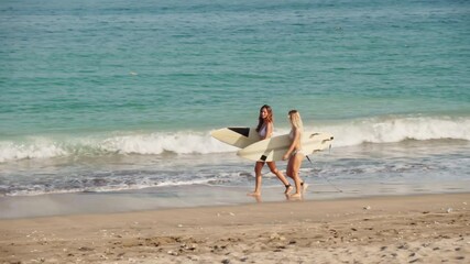 Sequence of shots of two joyful young women wearing white bikini swimsuits walking along beach holding surfboards