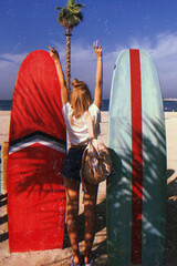 A young skinny girl on the beach standing between two big bright red and blue surfing boards seen from behind with her arms high above the head showing peace sign
