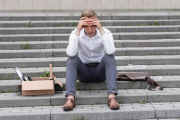 A fired office worker sits on the steps. The man does not know what to do next. Next to it is a cardboard box with stationery.
