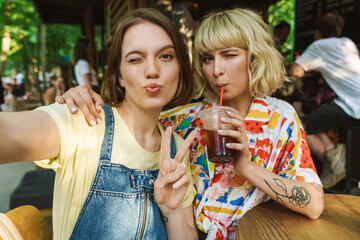 Image of women taking selfie and gesturing peace sign while drinking soda