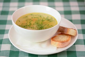 Chicken soup with vegetables and meat served in a white bowl over rustic wooden background with green plaid tablecloth.