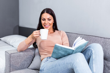Young woman at home sitting on modern sofa relaxing in her living room reading book and drinking coffee or tea. Cozy bed and a beautiful girl, reading a book, concepts of home and comfort