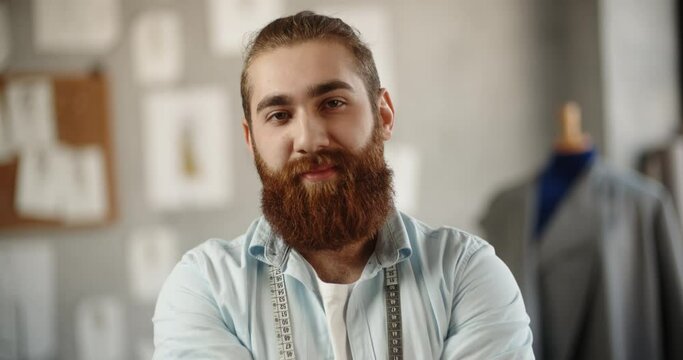 Close-up portrait shot of bearded hipster clothing designer working in his office, looking at camera and positively smiling - small business, fashion concept 4k footage