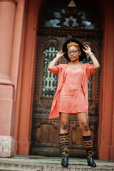 African red haired woman wear eyeglasses, orange dress and black hat posing outdoor.