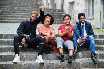 Fototapeta premium Four african friends having fun outdoors. Two black girls with guys sit on stairs of an old city.