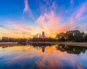 Fototapeta na wymiar Dawn over Victory Park on Poklonnaya Hill in Moscow in the summer. Colorful blue sky with clouds and sunrise on the horizon above an Orthodox church. The sky is reflected in the pool of the fountain.