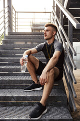 Image of unshaven sportsman drinking water while sitting on stair