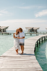 Happy loving couple walking on summer tropical beach on wooden jetty with overwater wooden bungalow on background