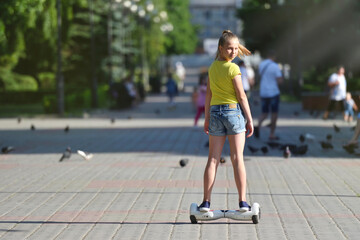 A girl child riding a hoverboard in a park and turning back and looking at the camera on a summer day