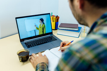 Back view of crop unrecognizable busy male in casual clothes taking notes in notebook while watching video presentation of female colleague on laptop during online conference via laptop