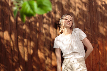 Portrait of a pretty girl in wireless headphones, standing against a wooden fence in the backyard on a Sunny day.