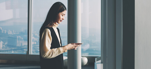 Young Asian woman is reading text message in network via smart phone,while is standing in modern interior with cityscape New York view.Female office worker of successful company is using mobile device