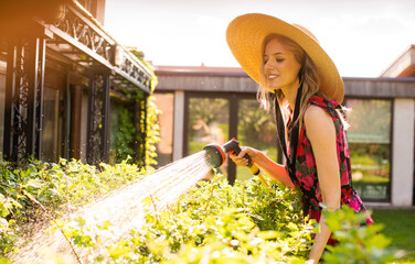 Pretty smiling girl in a hat watering plants with a garden hose in the garden in summer,...