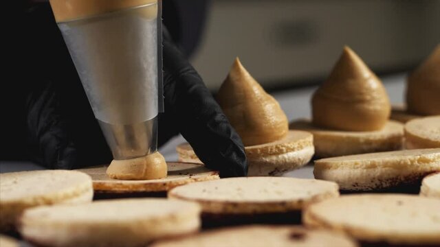 Chef is making caramel macarons. Hand with confectionery bag squeezing cream filling to macarons shells, close-up.